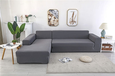 Funda elástica gris para sofá en forma de L en un salón moderno, complementada con decoraciones y planta verde, perfecta para renovar y proteger el mobiliario.