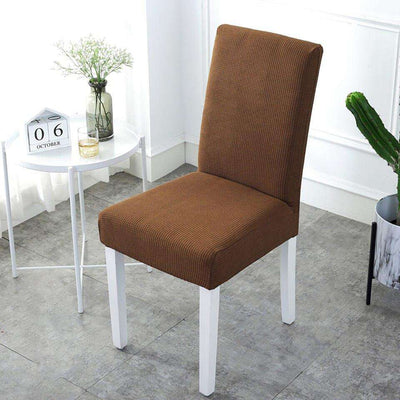 Funda de silla de color café claro ajustable, en un ambiente luminoso y moderno con mesa blanca y decoración minimalista, ideal para renovar el mobiliario de hogar o oficina. Disponible en blueemoonco.com.