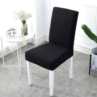 Funda de silla elástica negra en una silla blanca, ideal para decoración del hogar, vista en un ambiente minimalista con una mesa blanca, un florero y detalles decorativos, disponible en blueemoonco.com
