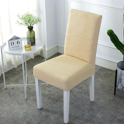 funda de silla beige ajustable en una silla blanca en una habitación luminosa con mesa blanca, planta verde y ventana con cortinas blancas, perfecta para decorar el hogar, disponible en blueemoonco.com