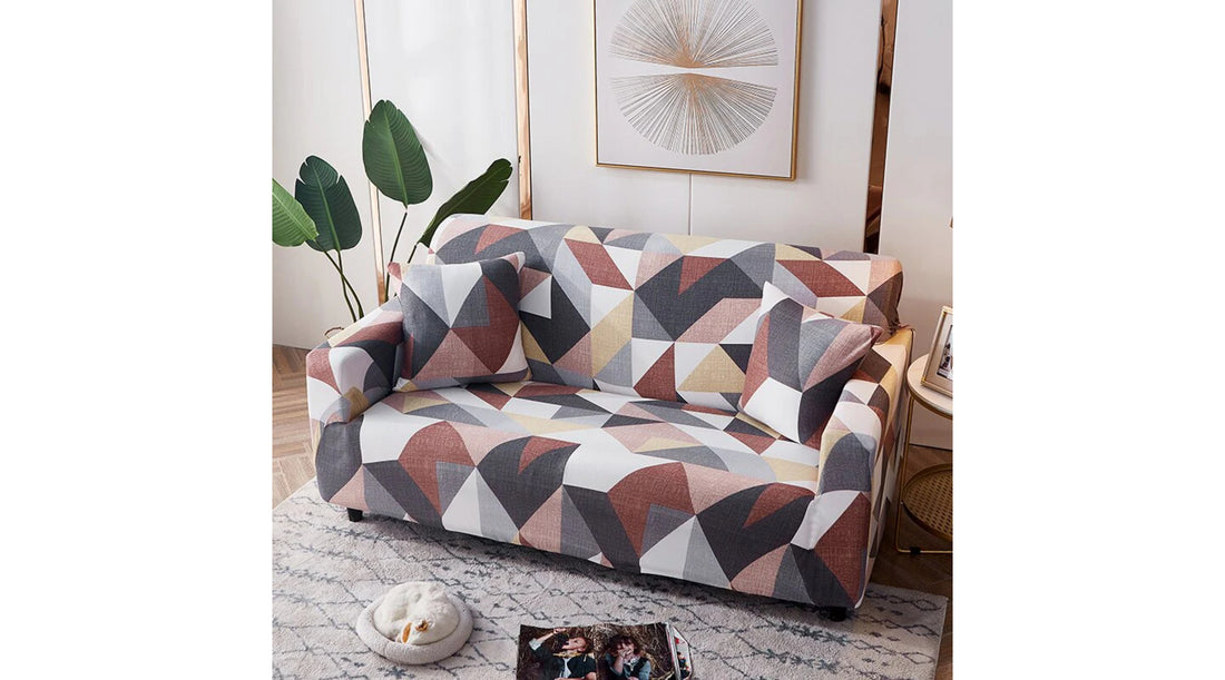 Sofá cubierto con una funda geométrica de colores cálidos, acompañado de cojines a juego, en una sala de estar moderna con decoración minimalista y plantas de interior.