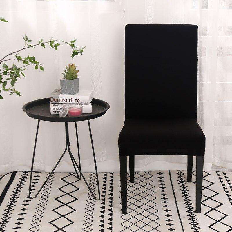 Funda universal de silla en color negro, estilo moderno con mesa auxiliar negra con libros, vela y planta pequeña, sobre una alfombra estampada y cortina traslúcida al fondo. Ideal para decoración contemporánea. Disponible en blueemoonco.com