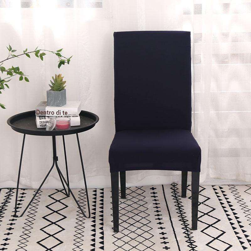 Funda de silla de color azul marino en tamaño universal, situada en un entorno moderno con una mesa lateral negra que sostiene libros y una planta decorativa, sobre una alfombra con patrones geométricos y trasfondo de cortina traslúcida