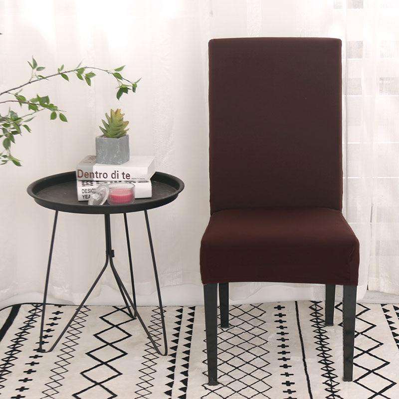 Fundas para sillas en color café oscuro de Blueemoonco.com, perfectas para un estilo moderno. Silla universal cubierta en una habitación con cortinas blancas, mesa auxiliar negra con libros y planta, sobre una alfombra con patrones geométricos.