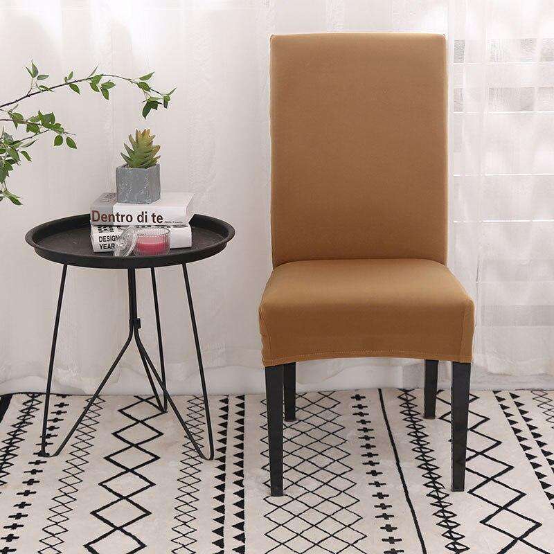 Funda de silla color camel en tela elástica y ajustable, presentada en un entorno moderno con mesa redonda negra, planta decorativa, libros y una alfombra geométrica, ideal para renovar el estilo de cualquier silla. Disponible en blueemoonco.com.