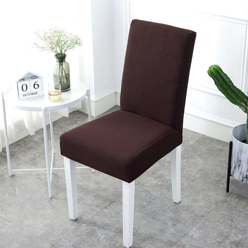 Fundas de silla color café oscuro, de diseño elegante y ajuste universal, en un entorno luminoso con mesa blanca y detalles decorativos verdes, perfectas para renovar el estilo de cualquier comedor o oficina, disponibles en blueemoonco.com